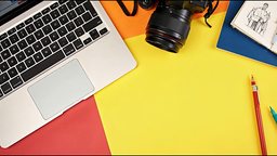 A flat lay of a laptop, camera, sketchbook and colored pencils