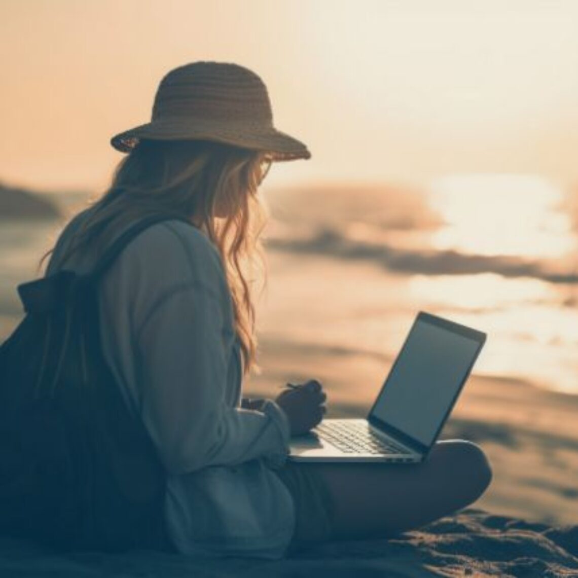 woman with long hair and wearing a hat sitting on a beach at dusk working on a her laptop